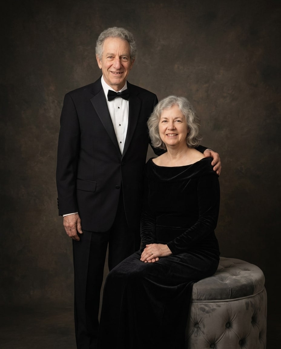 A formal grandparent memorial portrait featuring an elderly man in a black tuxedo standing beside a seated woman in an elegant black velvet dress against a warm, dark studio backdr