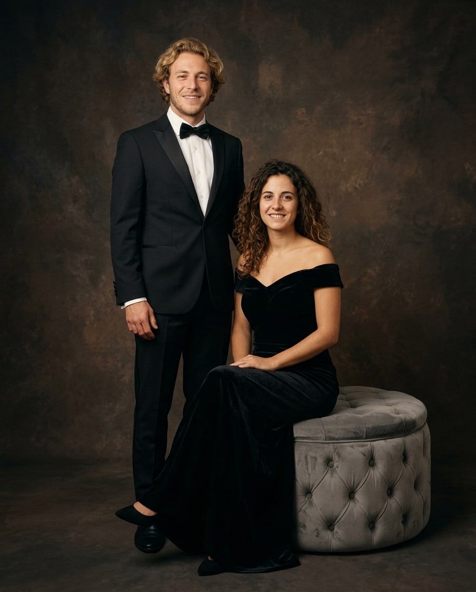 A man in a black tuxedo stands beside a woman in a black off-the-shoulder velvet gown seated on a grey tufted ottoman against a dark brown studio background.